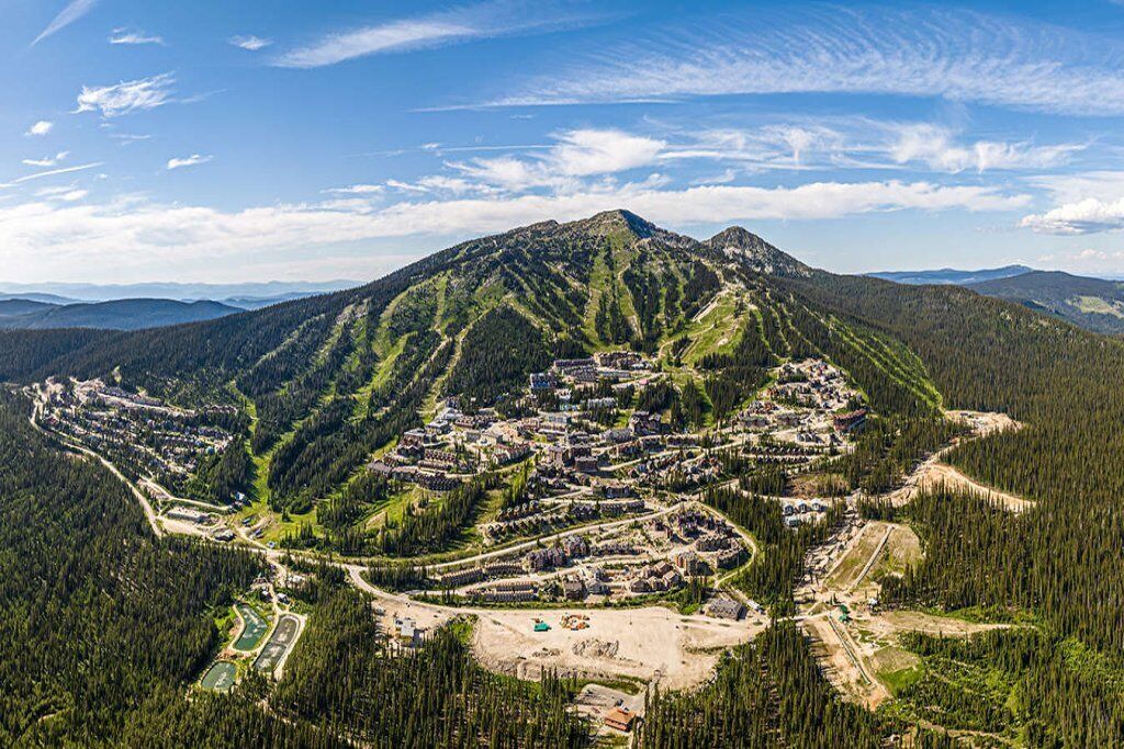 An aerial view of a mountain resort with a dense forest surrounding it, featuring a central cluster of buildings and a parking area.