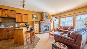 A cozy living room with a fireplace, a kitchen area with wooden cabinets, and a comfortable seating area with a leather sofa. - Bear's Paw Chalet - Amazing Location & Ski In/out (Beaverdell)