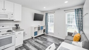 Une cuisine moderne avec des armoires blanches, un four à micro-ondes et une cuisinière. Le salon est équipé d’un canapé gris avec un coussin jaune et d’une table blanche.