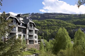 A multi-story hotel with a modern architectural design, featuring balconies and large windows, set against a backdrop of lush green hills and a clear blue sky. - Great Family ski location (Beaver Creek)