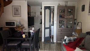A living room with a grey sofa, a dining table with chairs, a kitchen area with cabinets, and a microwave. - Maison au Calme à Proximité du lac (Hourtin)