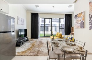 A modern living room with a dining area, a flat-screen TV, and a large window with a view of a building and greenery.