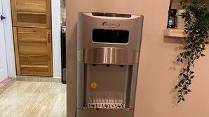 Ein Edelstahl-Wasserspender mit Bedienfeld und Wasserflaschenhalter, platziert in einer Küche mit Holzschränken und Fliesenboden.