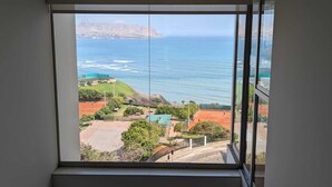 Blick aus einem Fenster auf einen Tennisplatz, ein Schwimmbad und eine Küstenlandschaft.
