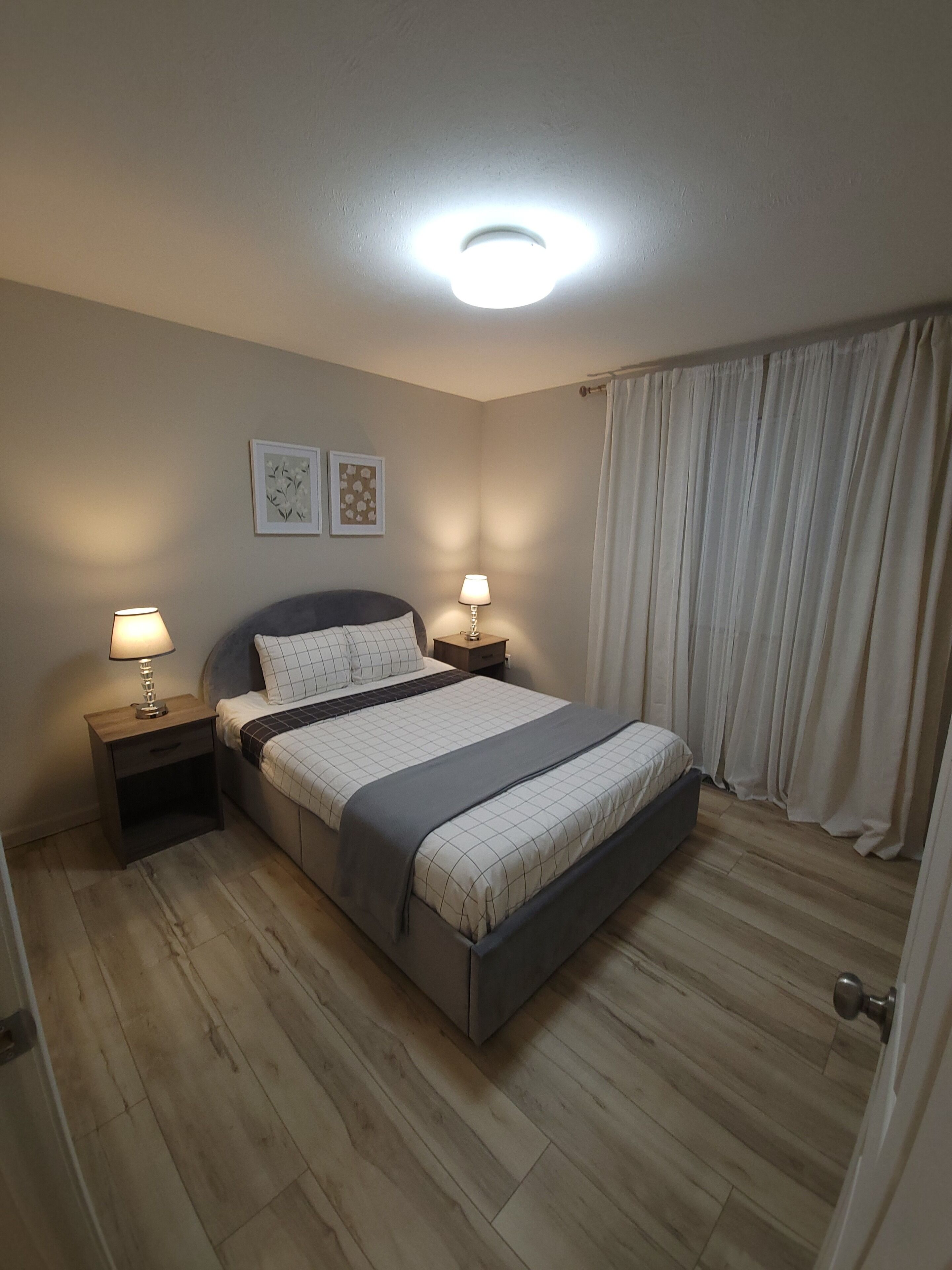 Ein Schlafzimmer mit einem Bett, zwei Nachttischen mit Lampen, einer Deckenleuchte und einem Fenster mit Vorhängen.