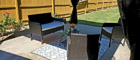 Een terras buiten met rieten meubilair, een kleine tafel met een bloemenvaas, en een kamerkleed met een patroon.