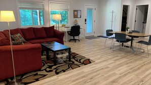 A modern living room with a red sofa, a coffee table, and a dining area with a table and chairs. There are windows with blinds, a lamp, and a door with a towel hanging on it. - Two Bedroom Relaxing Woodland Getaway with Putting Green and Walking Trails (Edwardsville)