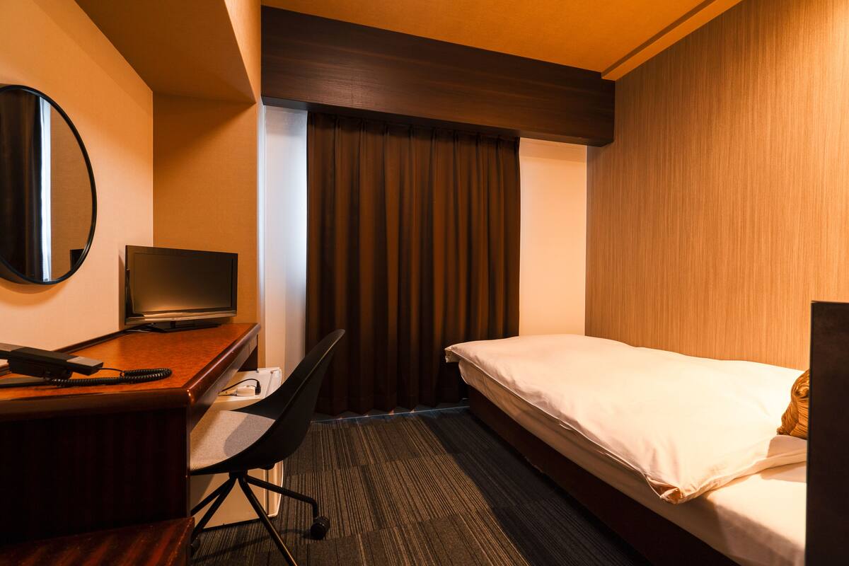 A hotel room with a bed, a desk with a computer, a chair, a TV, and a large window with curtains.