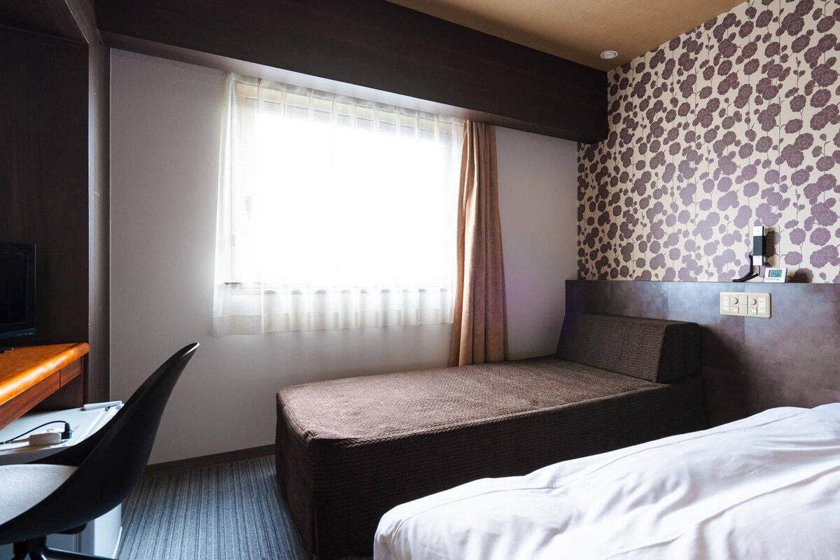 A hotel room with a bed, a desk with a chair, a window with curtains, and a wall with a floral pattern.