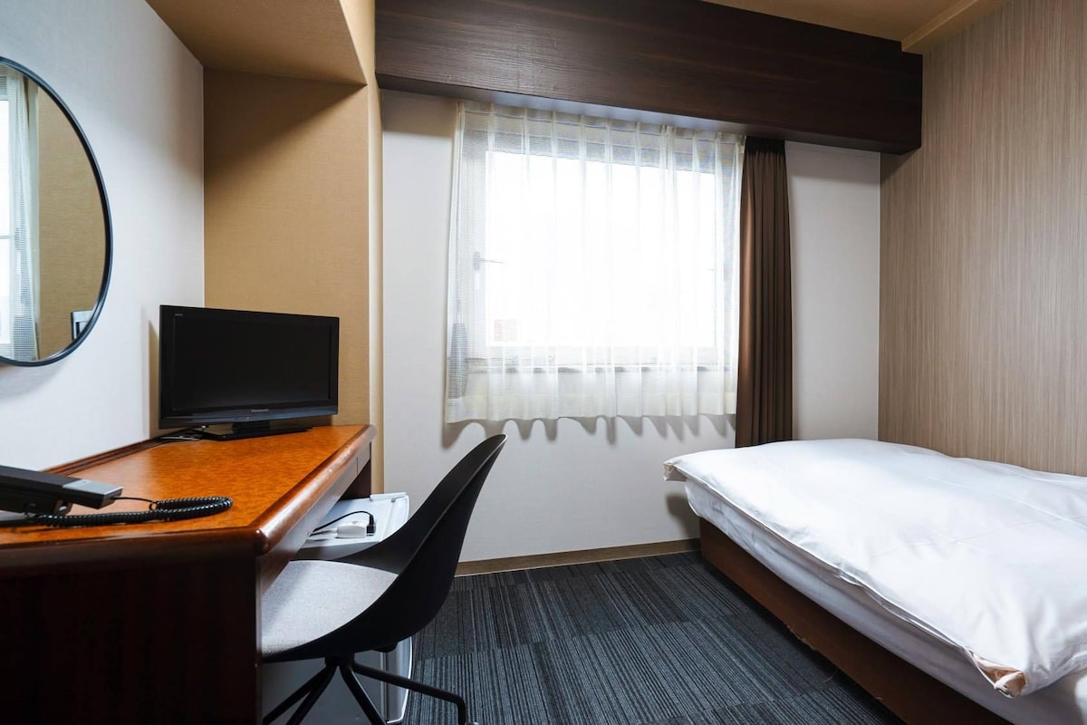 A hotel room with a bed, a desk with a computer, a chair, a window with curtains, and a mirror.