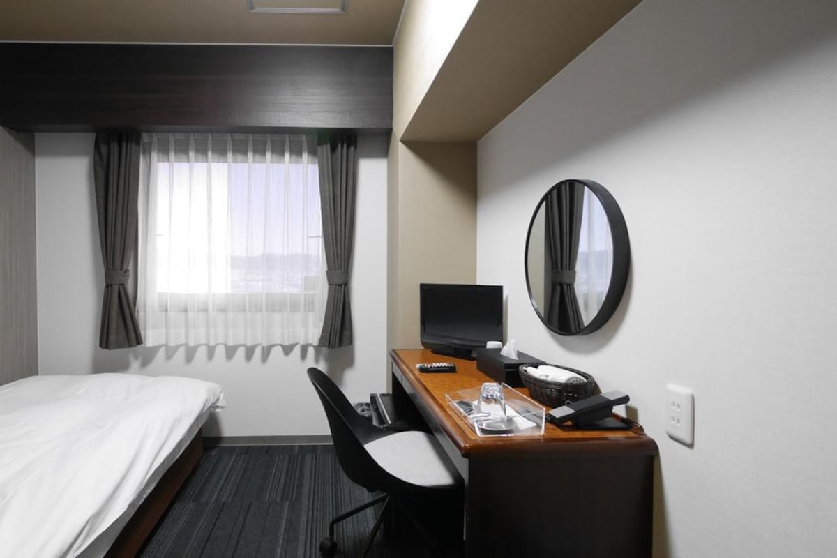 A hotel room with a bed, a desk with a computer, a chair, a desk lamp, a telephone, and a mirror on the wall.