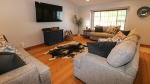 A living room with two sofas, a television, a wooden floor, and a window with blinds. - 5 Bedroom Lincoln Home 10 Minutes from Downtown (Lincoln)