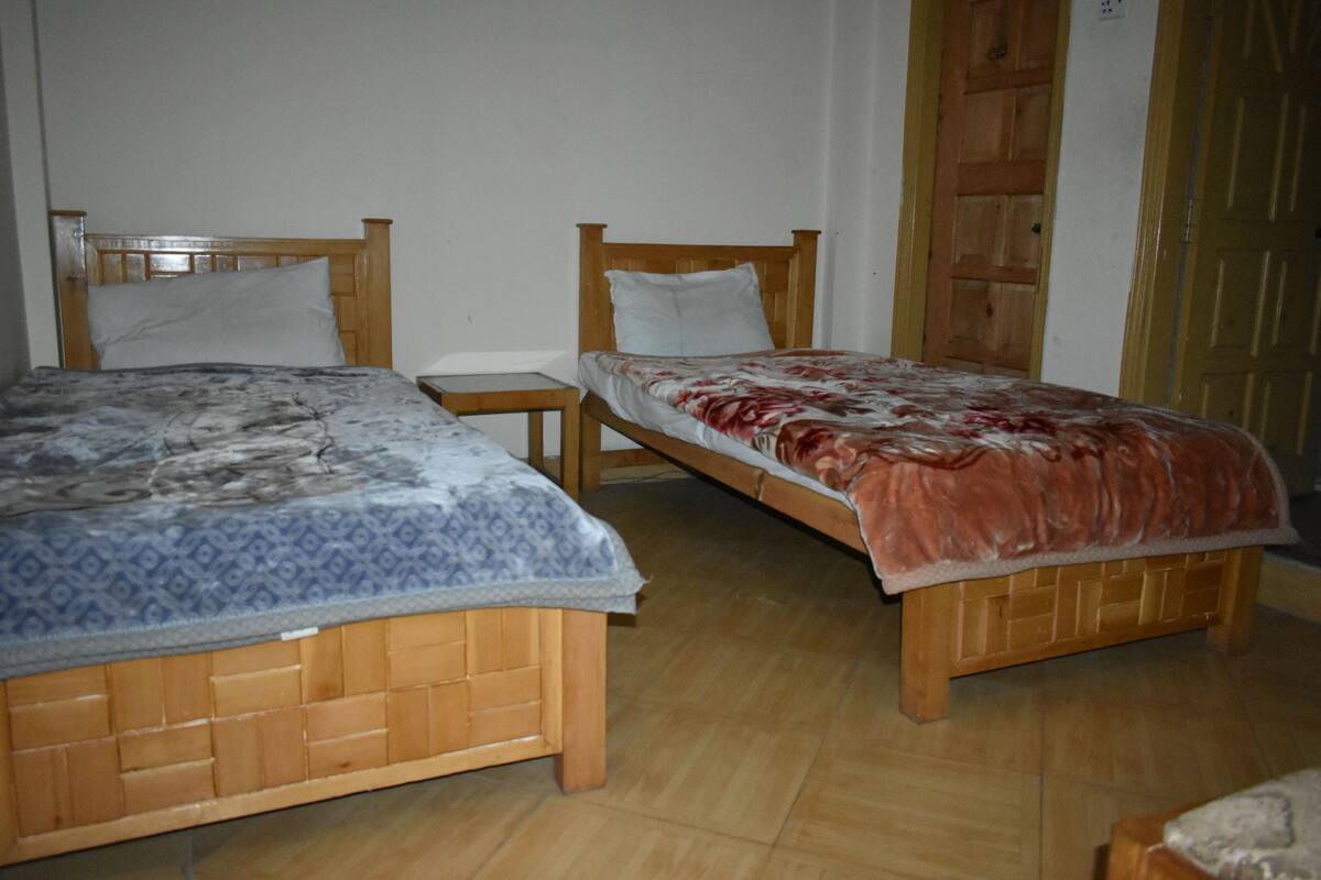 Une chambre avec deux lits en bois, chacun muni d’un oreiller et d’une couverture. Une table de chevet en bois se trouve entre les deux lits.