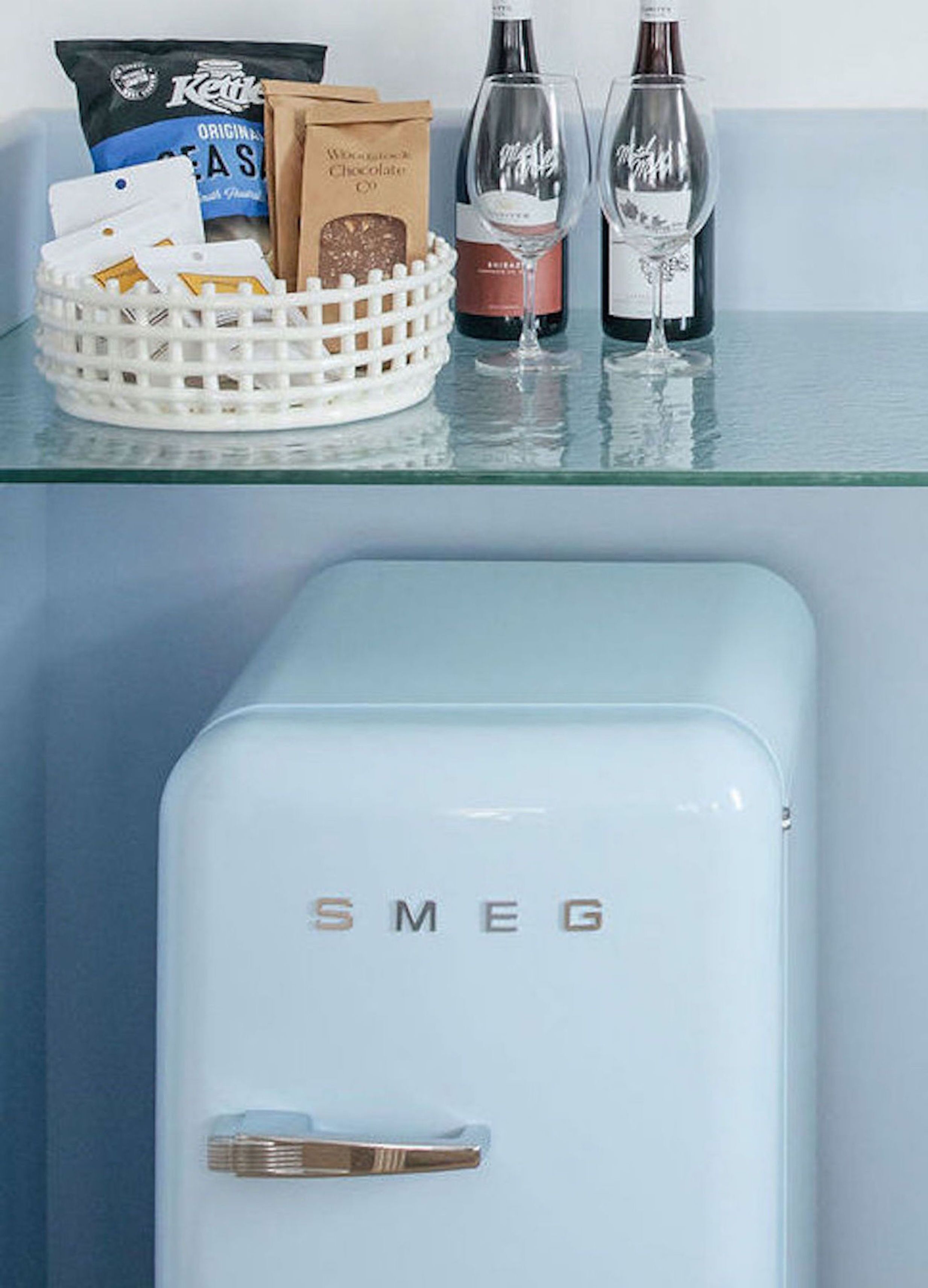 A kitchen counter with a white basket containing chocolate bars, a bag of coffee, and two wine glasses. Below, there is a light blue Smeg refrigerator.