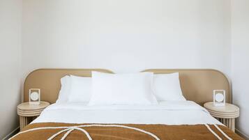 Una cama bien tendida con sábanas blancas y una manta marrón, dos mesitas de noche con altavoces, y una pared lisa.