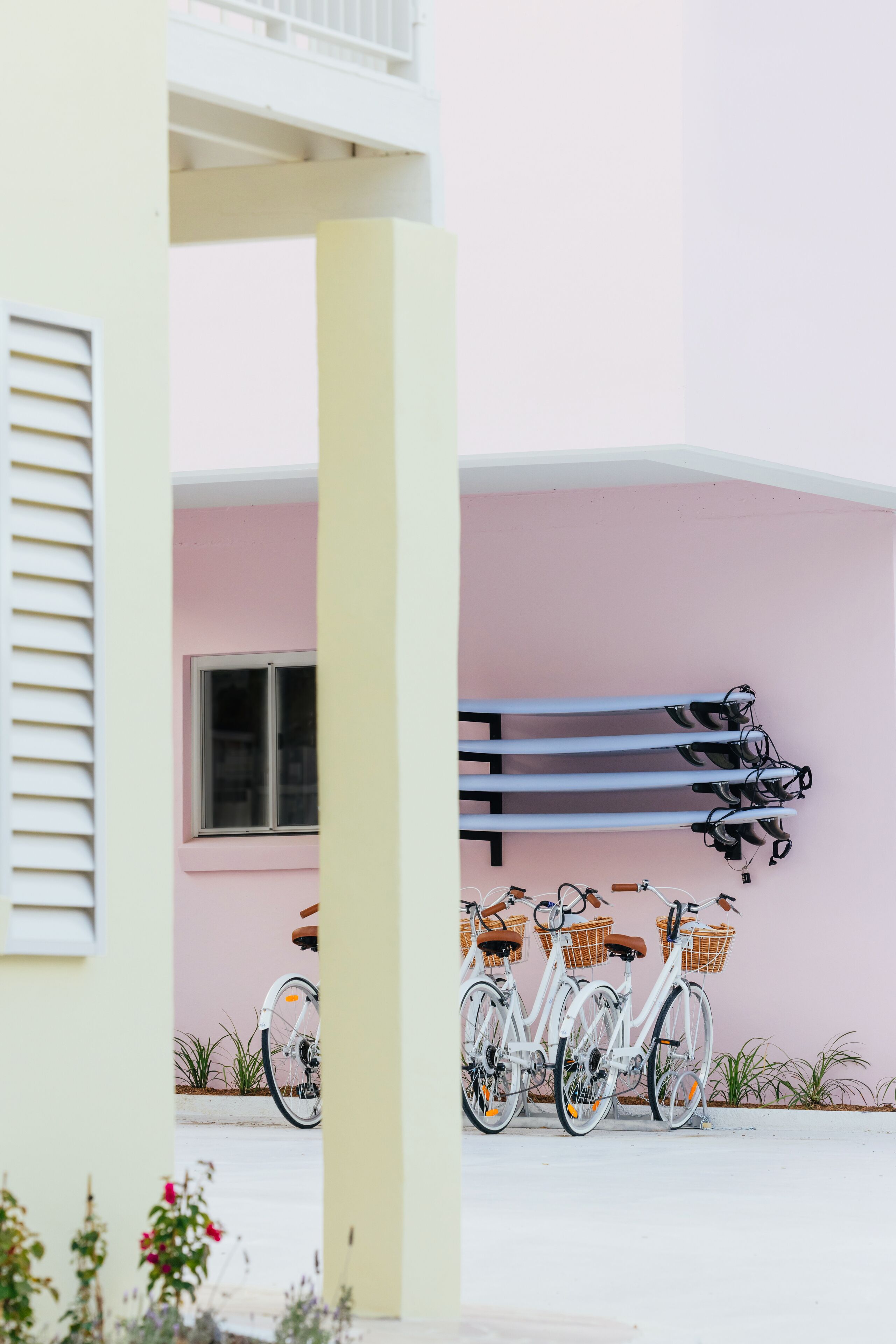 A building with a pink exterior, a white window, and a white door. There are three bicycles with baskets parked against the wall.