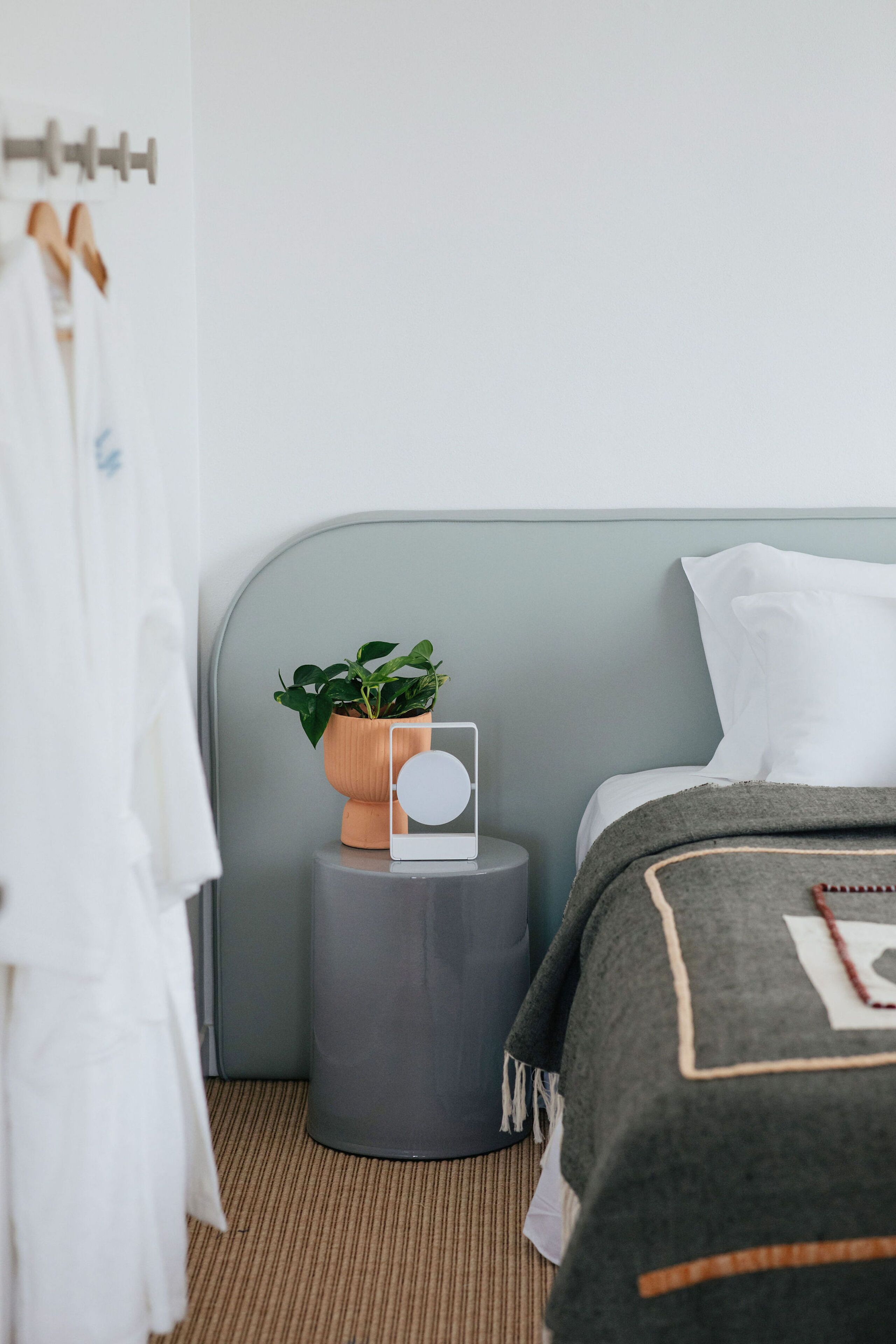 Un dormitorio ordenado con cama, mesita de noche con planta, espejo y bata colgada.