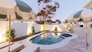 Piscina con sillas de descanso y sombrillas, rodeada de un muro blanco y plantas.