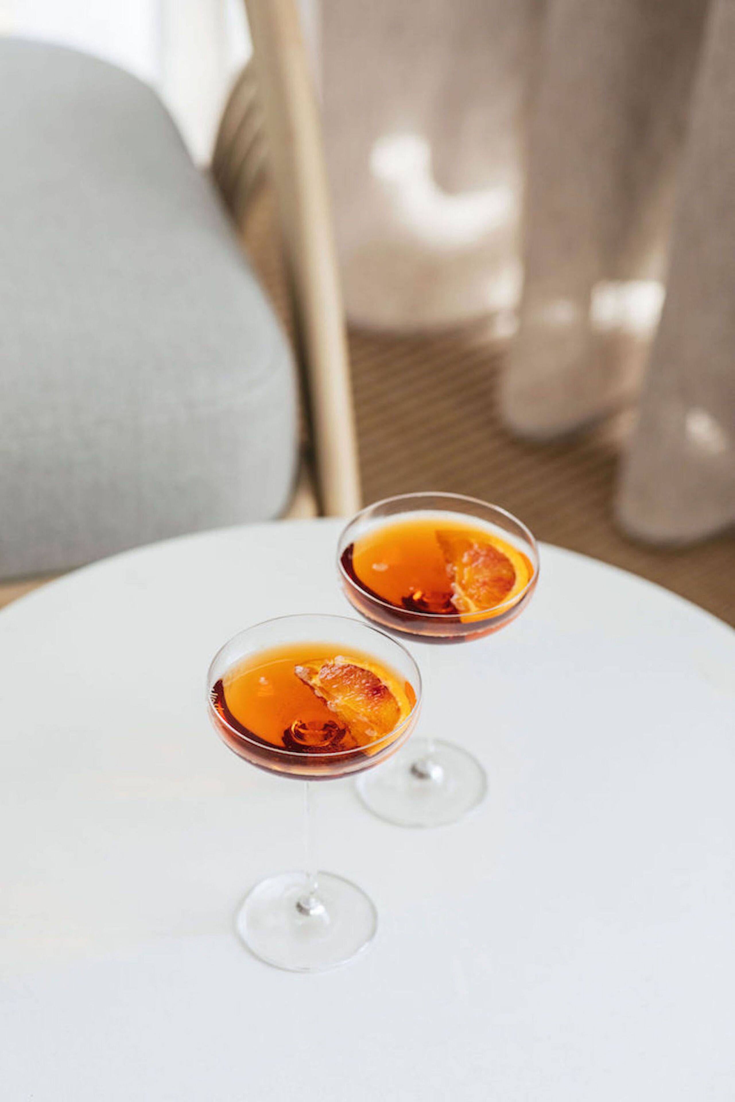 Two glasses of orange juice on a white table with a cushion in the background.