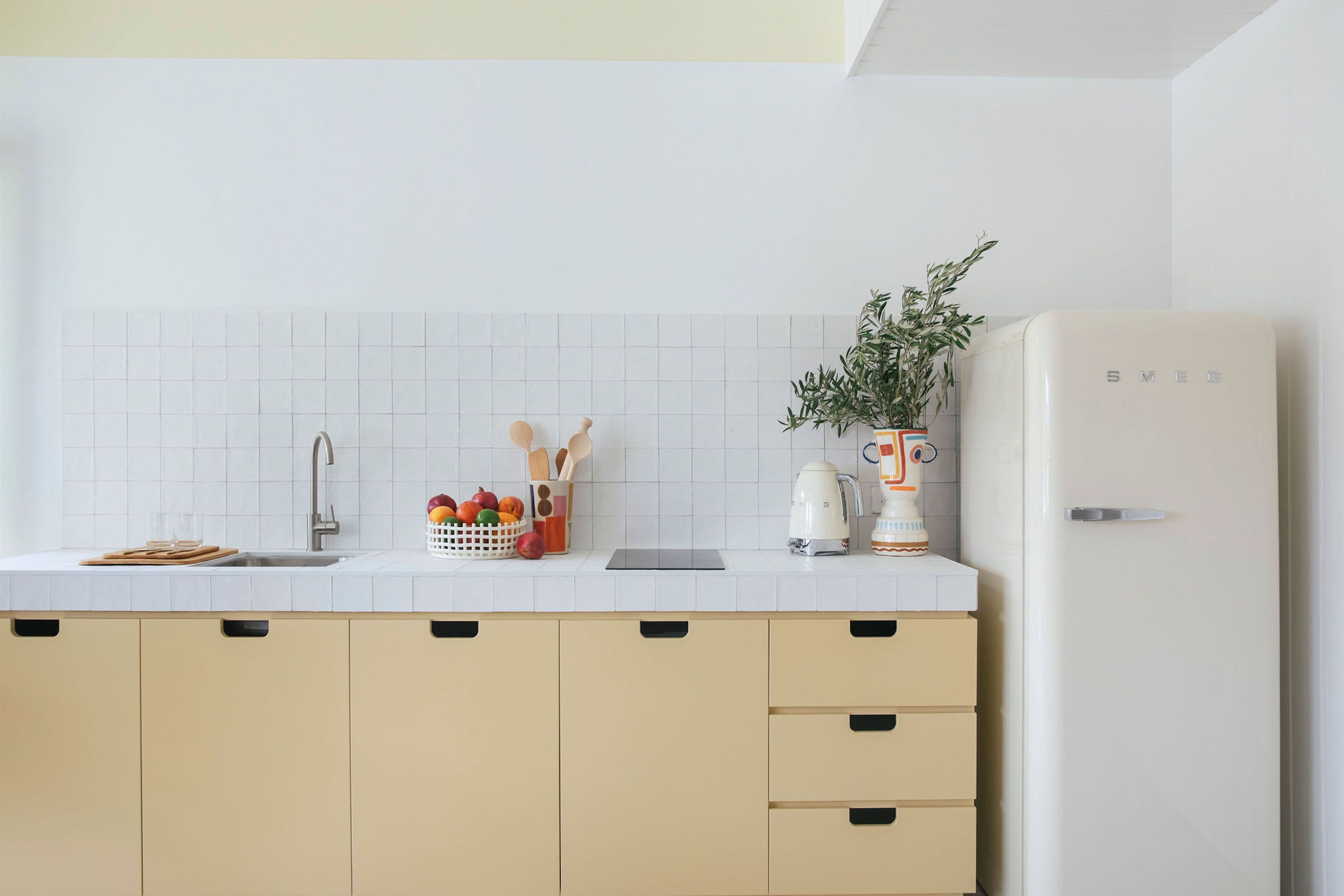 Una cocina moderna con salpicadero de azulejos blancos, un fregadero, una encimera con a cesta de frutas, una cafetera y un refrigerador.