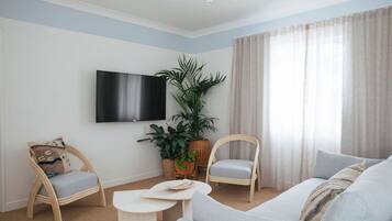 A living room with a flat-screen TV mounted on the wall, two wicker chairs, a grey sofa, a small round table, and a large potted plant.