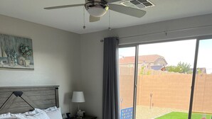 Ett sovrum med en säng, ett nattduksbord, en lampa, en takfläkt och ett stort fönster med utsikt över en trädgård.