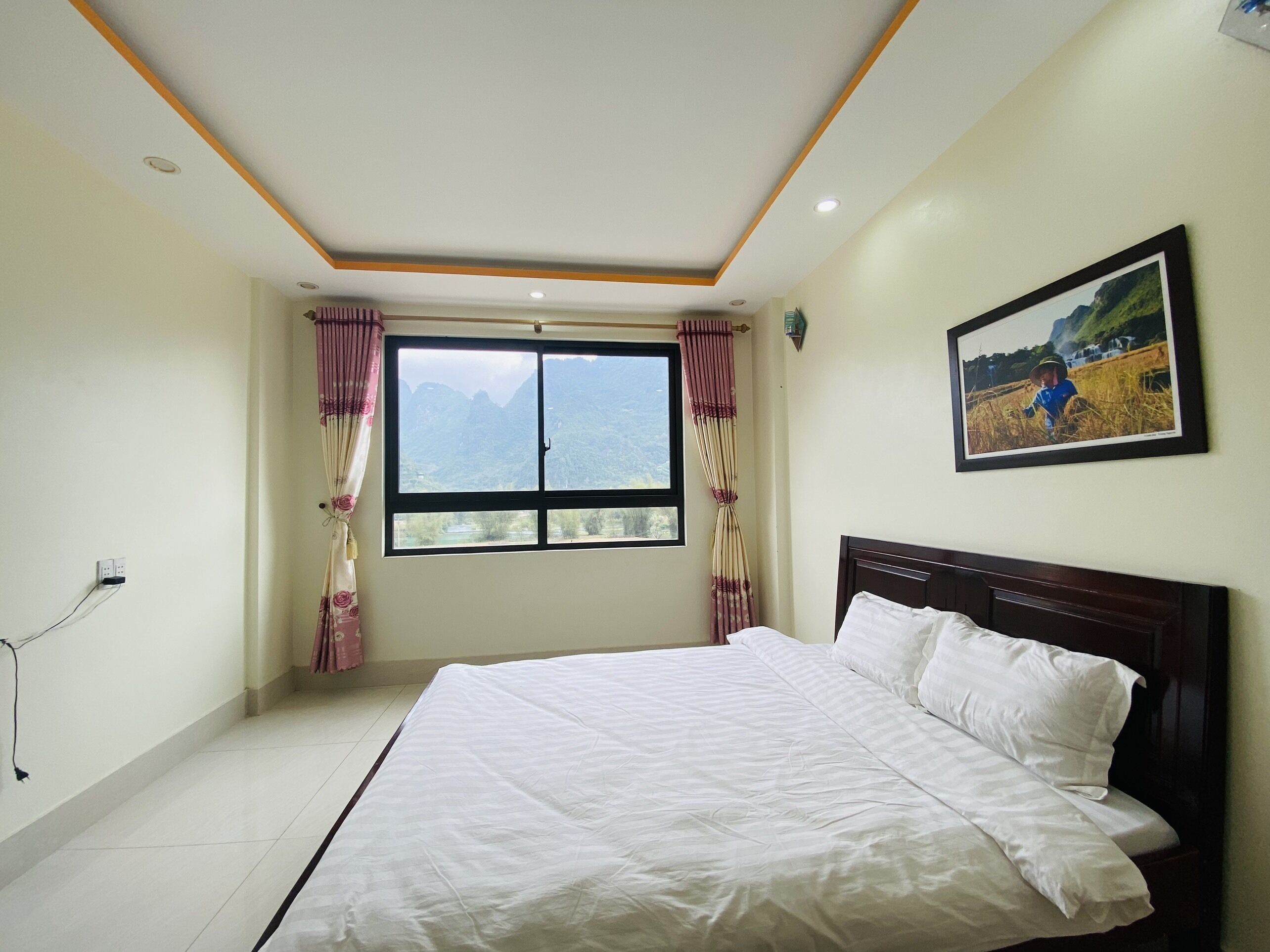 Un dormitorio con una cama grande, una ventana con cortinas, un cuadro colgado en la pared y un techo con iluminación empotrada.