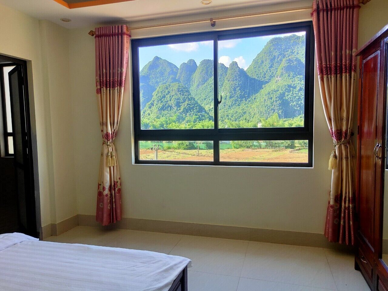 A room with a bed, a window with curtains, and a view of green mountains.