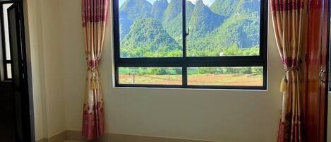A room with a bed, a window with curtains, and a view of green mountains.