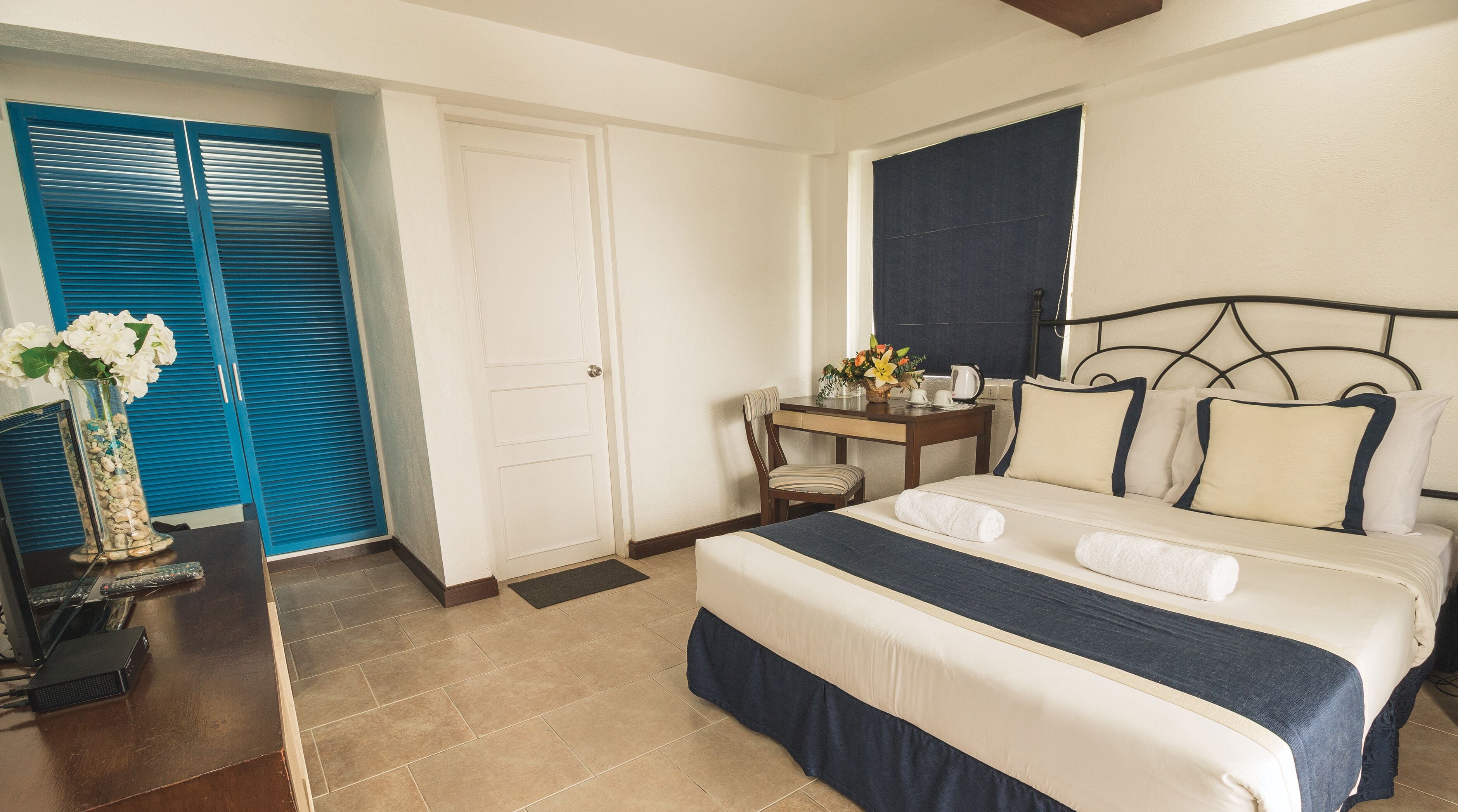 A hotel room with a bed, a desk, a chair, a vase with flowers, and a door with blue shutters.