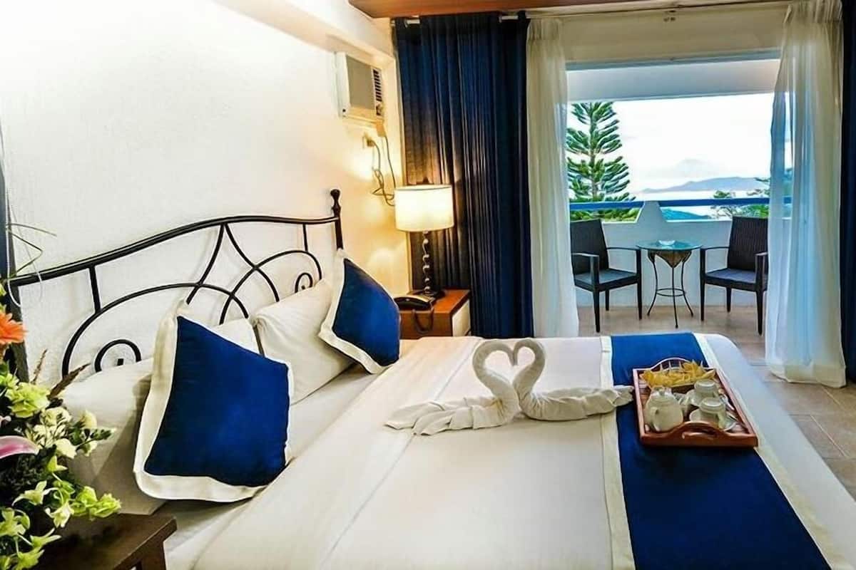 Une chambre d’hôtel avec un lit, un plateau contenant des articles pour le petit-déjeuner, un balcon avec une table et des chaises, et une vue sur la mer.
