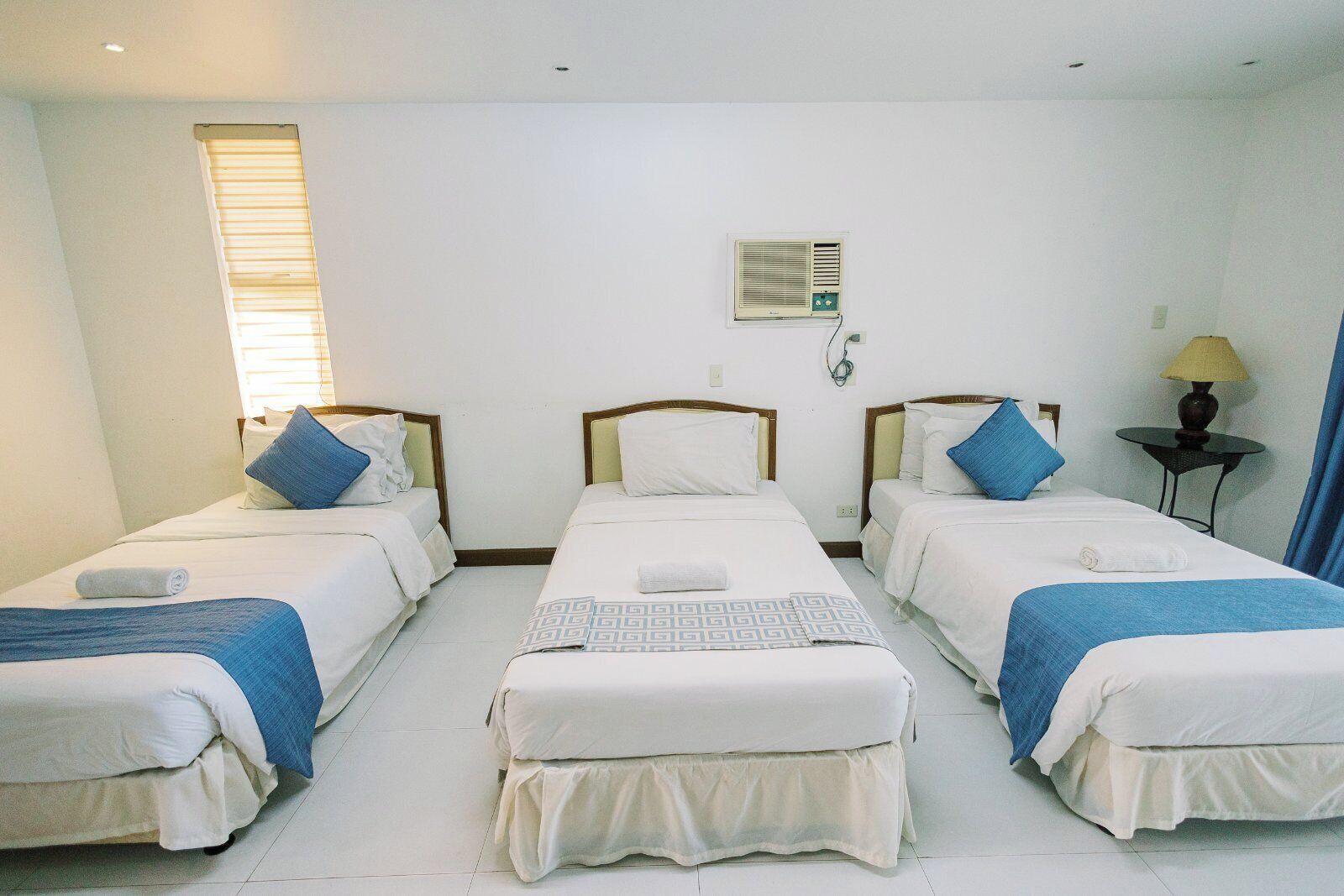 Une chambre d’hôtel avec trois lits simples, une petite table avec une lampe et une fenêtre avec des stores.