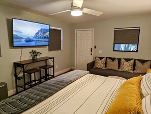 A bedroom with a bed, a TV mounted on the wall, a desk with a plant, a sofa with patterned cushions, and a window with a view.