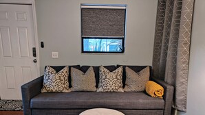 A living room with a grey sofa, patterned cushions, a small round ottoman, a window with a view, and a door. - King. Minutes to Wilton Drive. Private entrance and bathroom. (Wilton Manors)