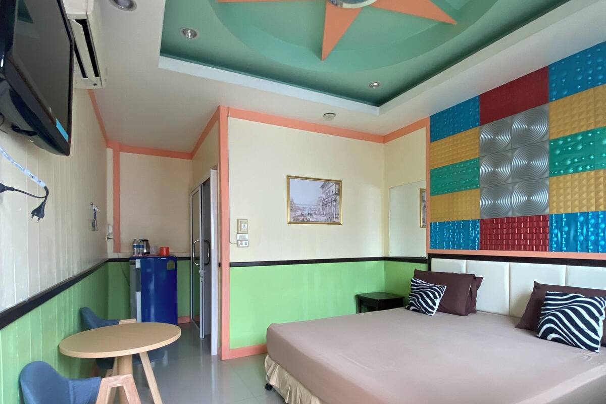 A hotel room with a bed, a small table, two chairs, a TV, and a colorful wall decoration.