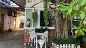 A small outdoor seating area with white chairs and a table, a tree with hanging plants, and a white picket fence. - Ruckunresort 1 (Nakhon Sawan)