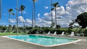 Une piscine à l’eau d’un bleu cristallin, entourée de chaises longues blanches et de palmiers, sous un ciel partiellement nuageux.