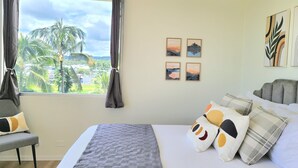 Une chambre à coucher avec un lit, des oreillers, une chaise, une fenêtre avec des rideaux et des œuvres d’art murales.