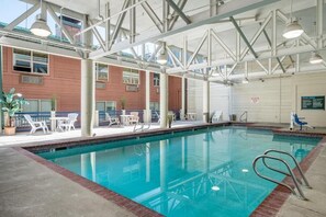 Uma piscina interior com água azul cristalina, rodeada por espreguiçadeiras e mesas brancas, com um edifício de tijolos ao fundo.