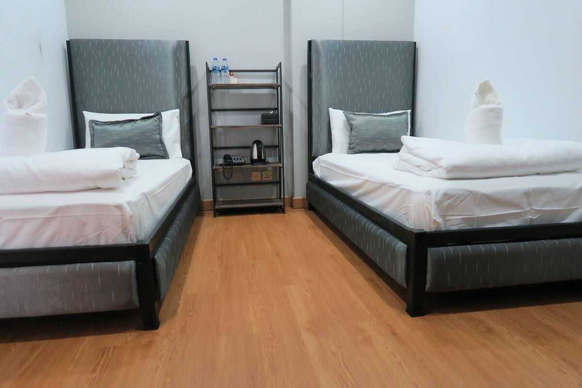 A hotel room with two single beds, a bedside table with a phone and a kettle, and a shelf with toiletries.