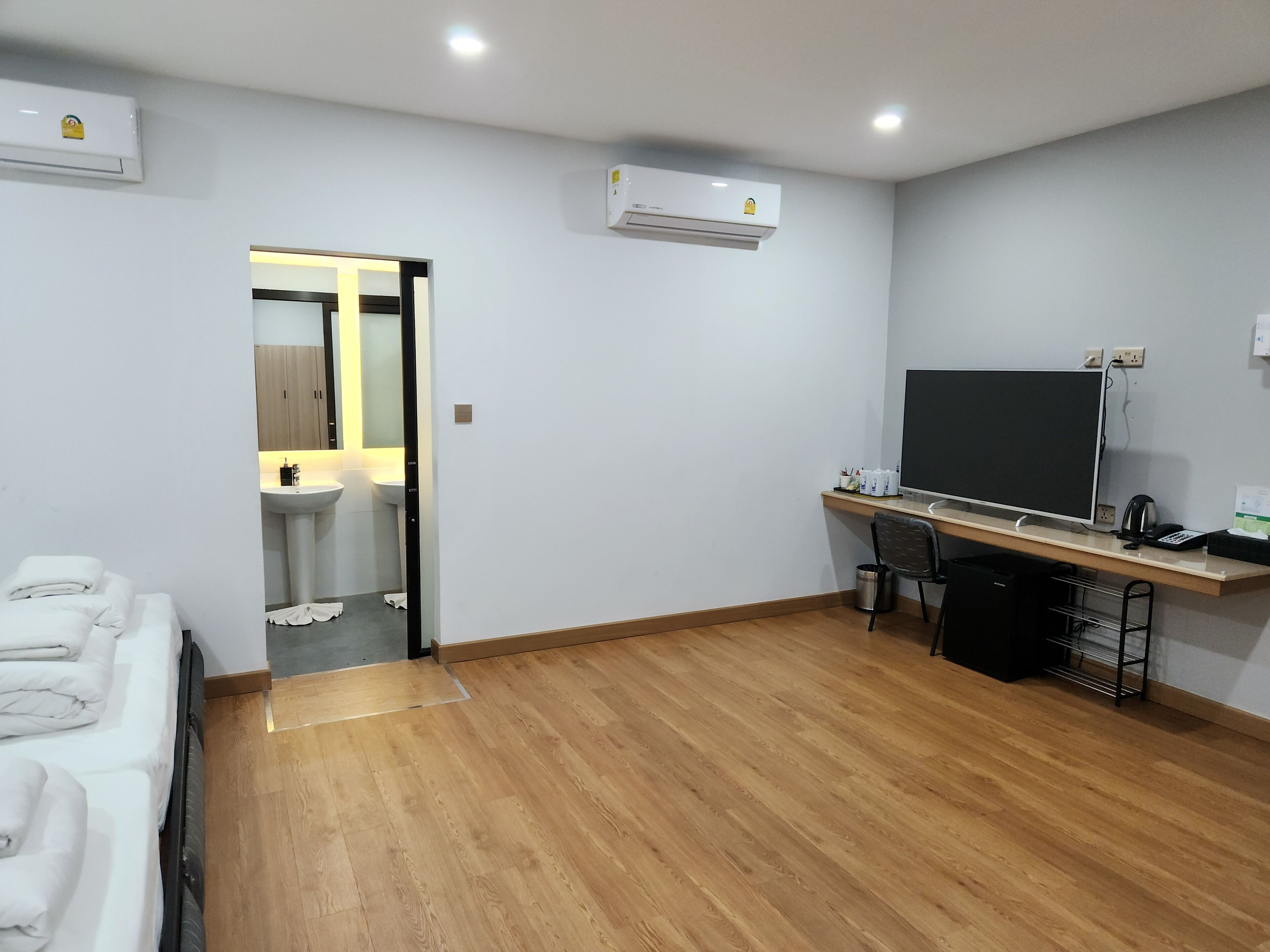Quarto de hotel moderno com piso de madeira, uma mesa com computador e TV de tela plana, cama com roupa de cama branca e um aparelho de ar condicionado na parede.