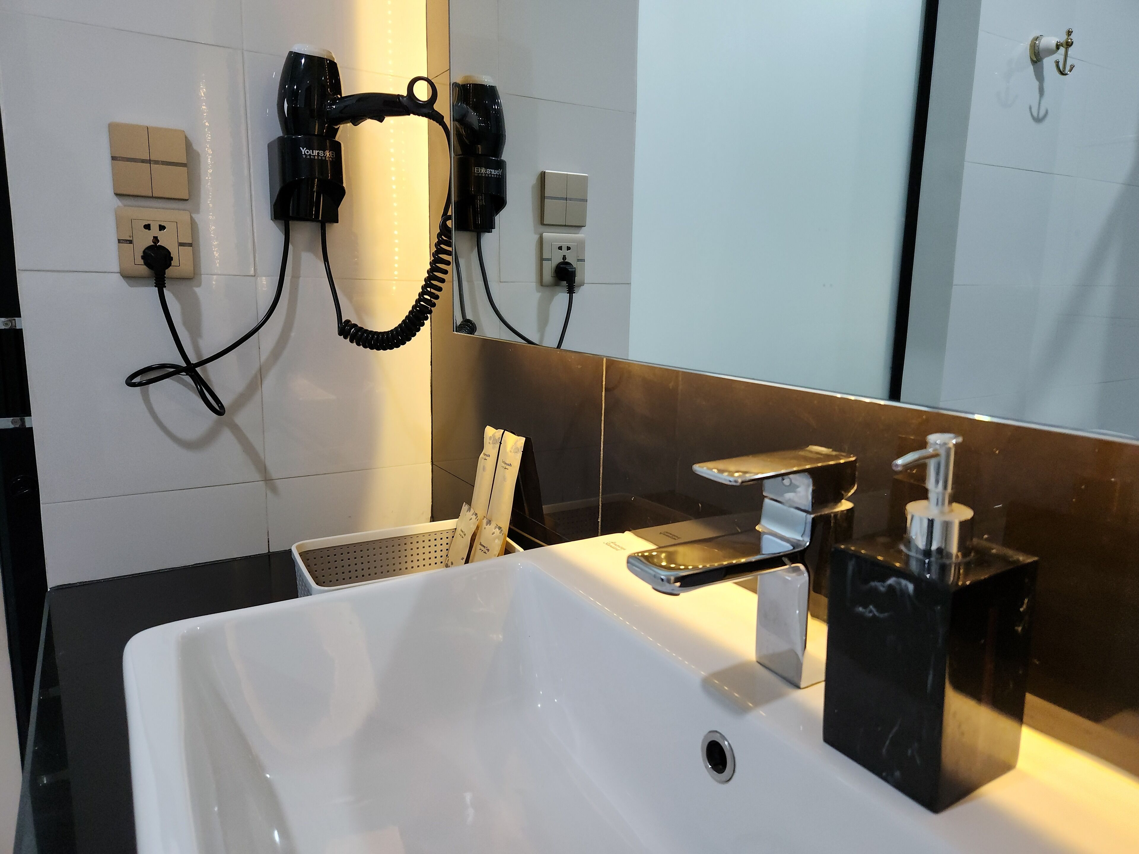 Un bagno moderno con un lavandino bianco, un piano di lavoro nero e uno specchio. Sono presenti due asciugacapelli montati a parete e un cestino portarifiuti.