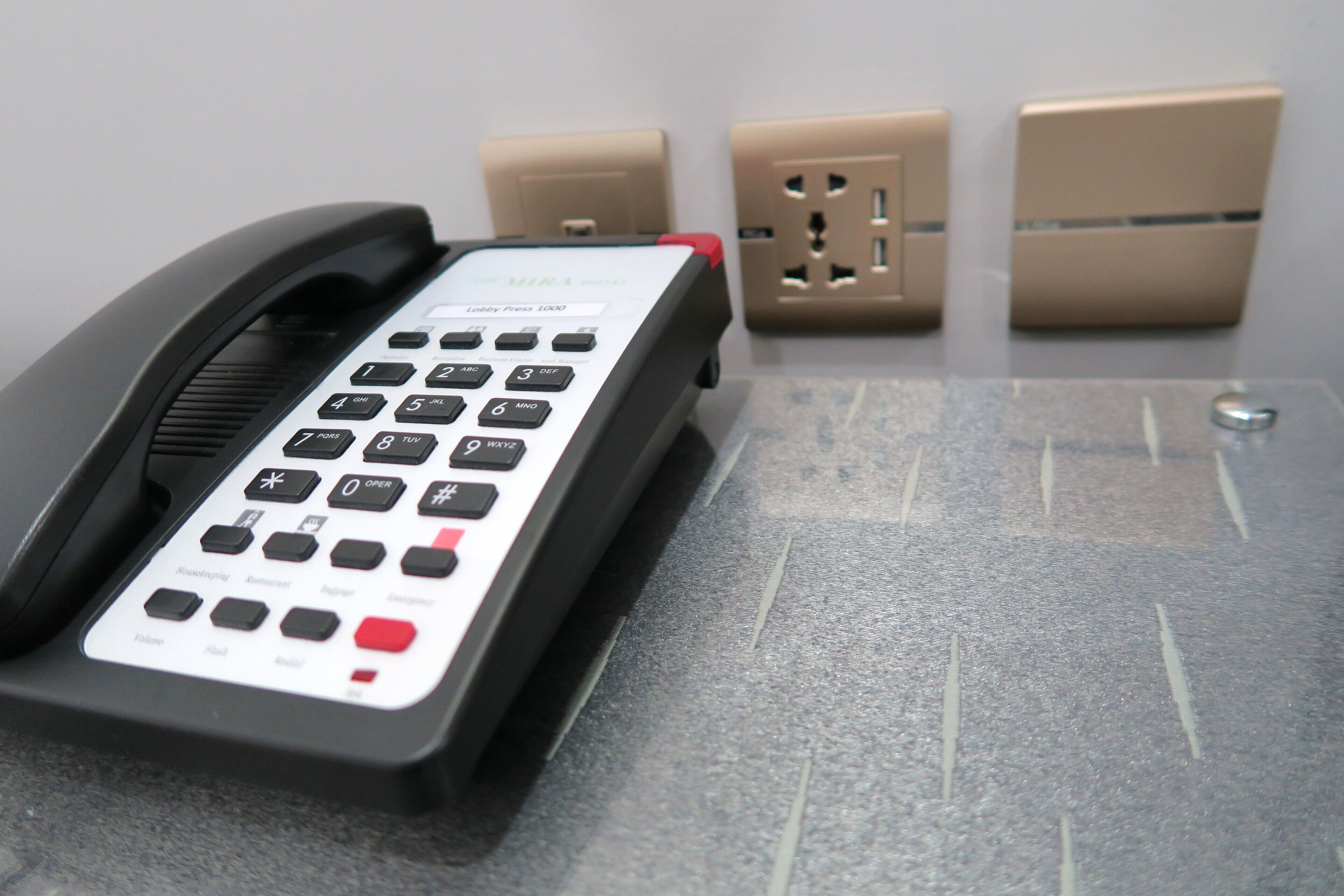A telephone with a keypad and a cord, placed on a textured surface with electrical outlets in the background.