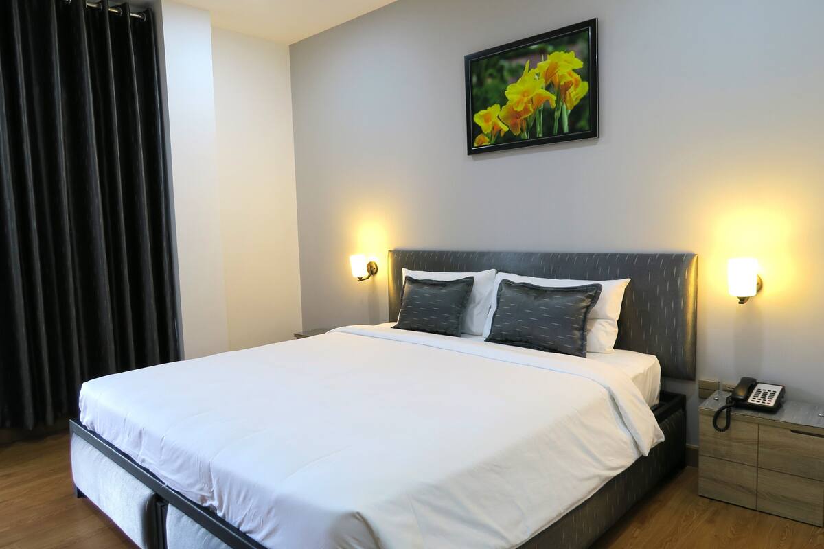 A neatly made bed with white linens, a headboard with a light wood finish, two bedside lamps, a telephone on a bedside table, and a framed picture of yellow flowers on the wall.