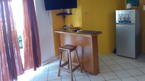 Une petite cuisine équipée d’un téléviseur fixé au-dessus d’un comptoir, d’un réfrigérateur et d’un tabouret en bois.