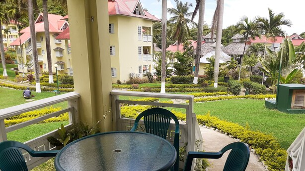 Une terrasse avec une table ronde et quatre chaises, donnant sur un jardin bien entretenu, agrémenté d’arbustes taillés et de palmiers.