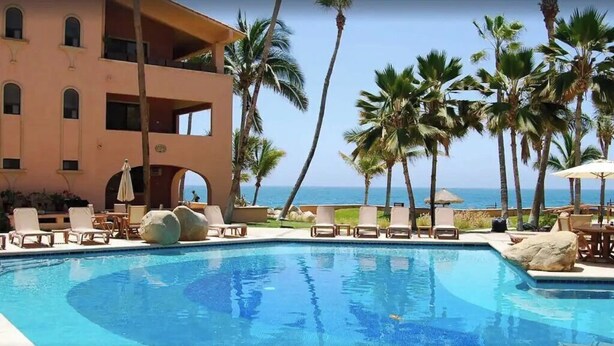 Un espace piscine aménagé avec des chaises longues, des palmiers et un bâtiment doté de balcons offrant une vue sur l’océan.