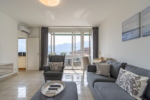 A modern living room with a grey sofa, a grey armchair, and a coffee table with remote controls. A dining area with a table and chairs is visible through a glass door, leading to a balcony with a view of mountains. - Walking distance to the Station with mountain and city views (Lugano)