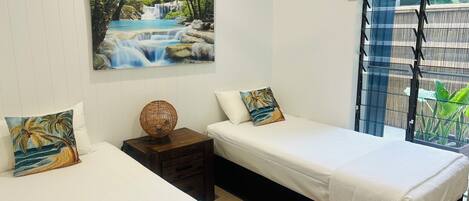 A bedroom with two beds, a nightstand, a wall-mounted lamp, and a window with a view of greenery.