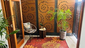 A room with a wooden door, a chair, a small table, a potted plant, and a decorative panel with circular patterns. -  Murriyan A Tropical Villa (Port Douglas)